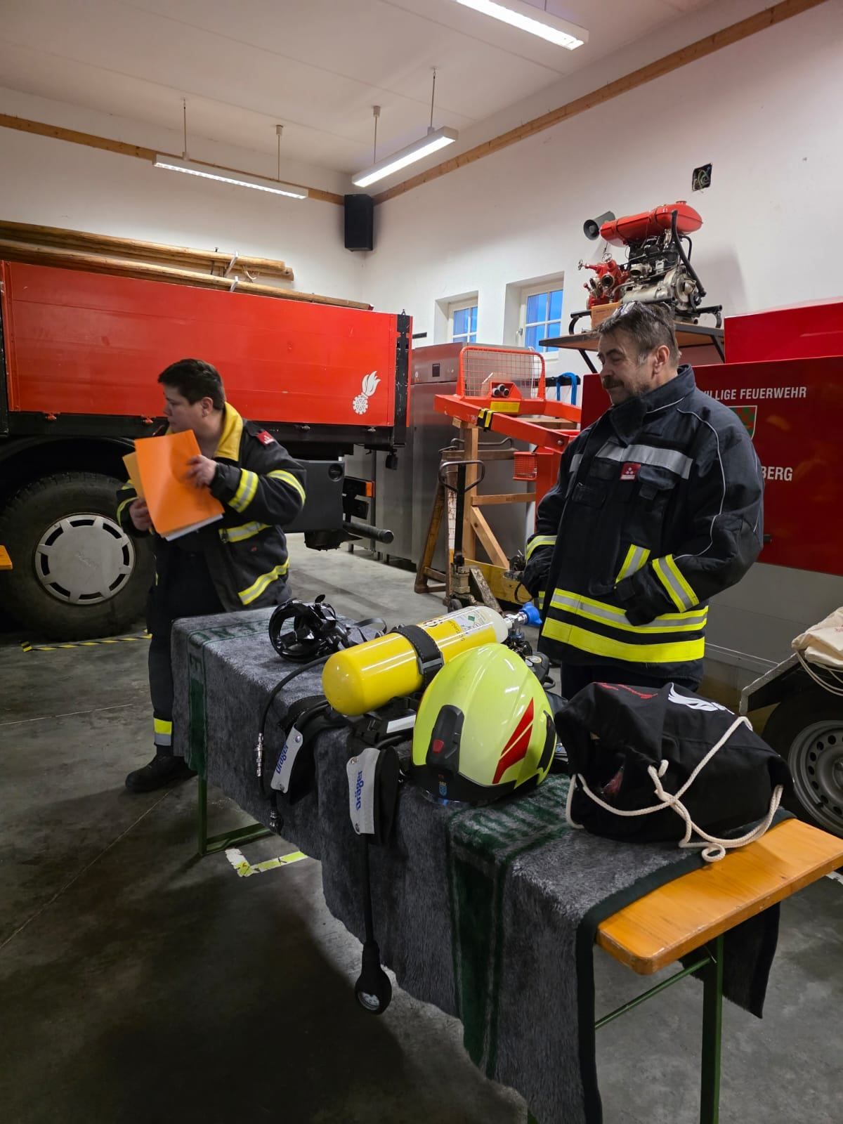 Zwei Feuerwehrleute stehen in der Garage eines Feuerwehrhauses. Ein Feuerwehrwagen ist hinter ihnen geparkt. Auf dem Tisch befinden sich verschiedene Ausrüstungsgegenstände, darunter ein gelber Helm und eine schwarze Tasche.