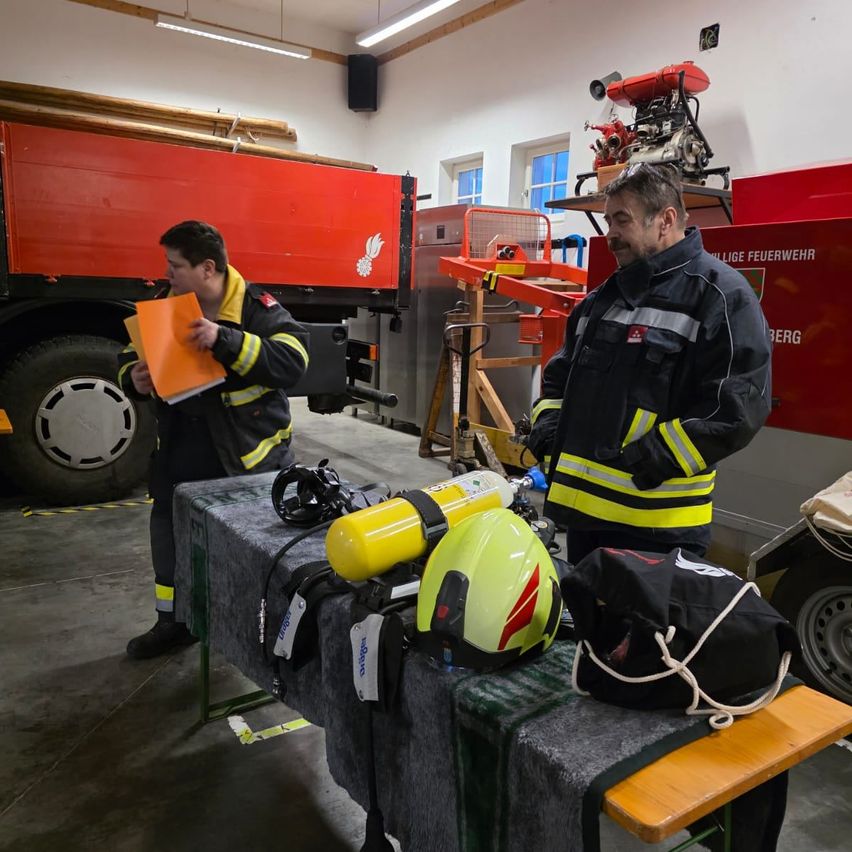 Zwei Feuerwehrleute stehen in der Garage eines Feuerwehrhauses. Ein Feuerwehrwagen ist hinter ihnen geparkt. Auf dem Tisch befinden sich verschiedene Ausrüstungsgegenstände, darunter ein gelber Helm und eine schwarze Tasche.