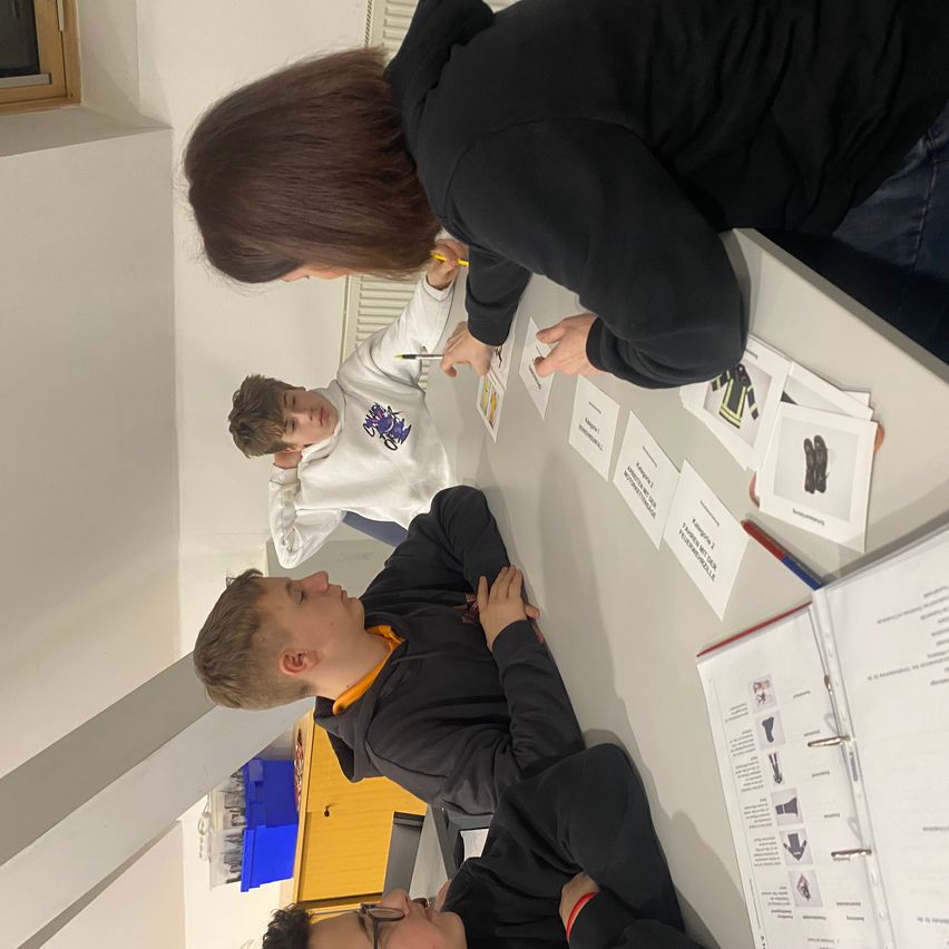 Drei Jungen und eine Frau sitzen um einen Tisch mit Papieren und Schreibutensilien. Die Frau schreibt auf einem Papier, während die Jungen zusehen.