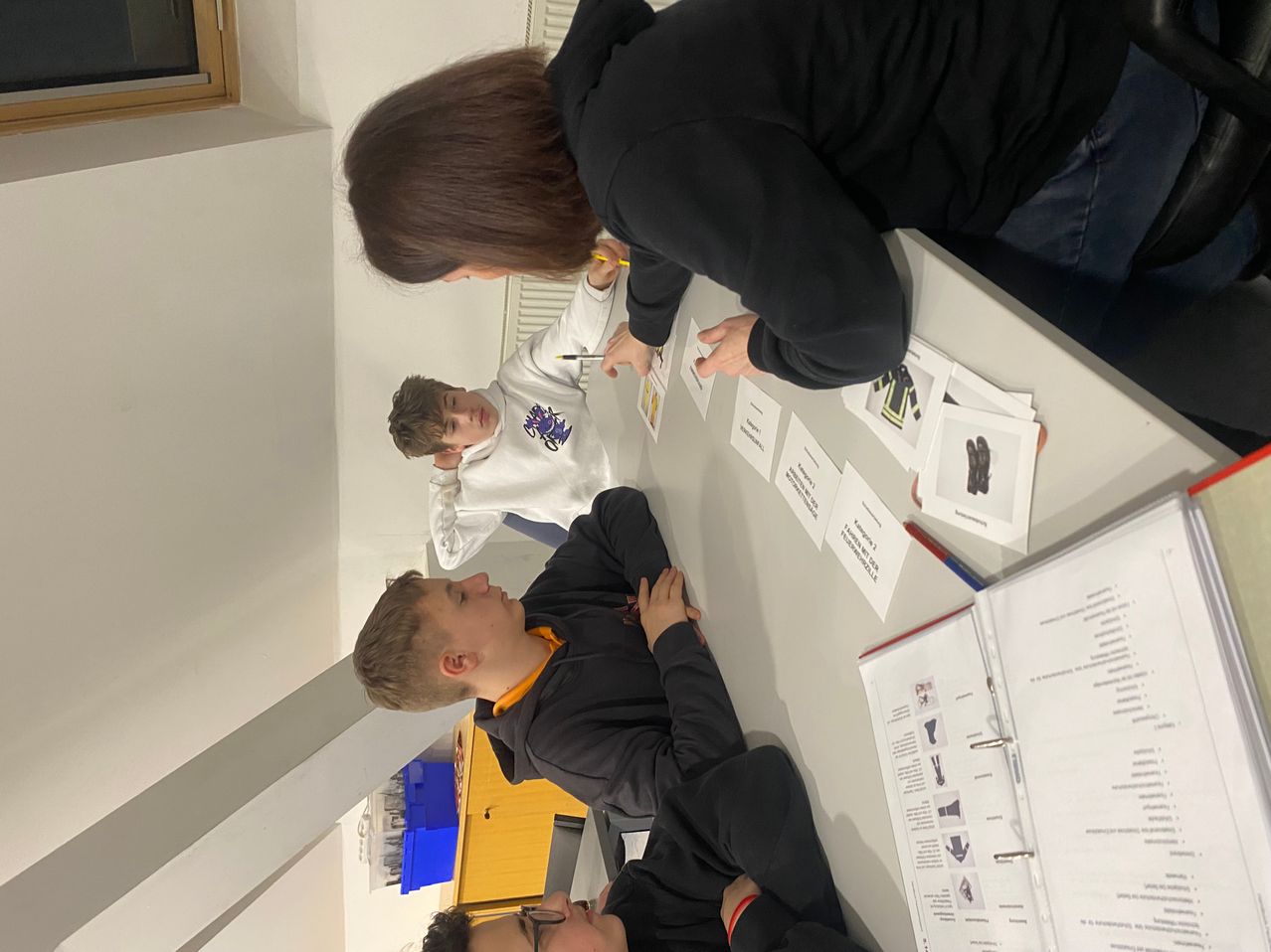 Drei Jungen und eine Frau sitzen um einen Tisch mit Papieren und Schreibutensilien. Die Frau schreibt auf einem Papier, während die Jungen zusehen.