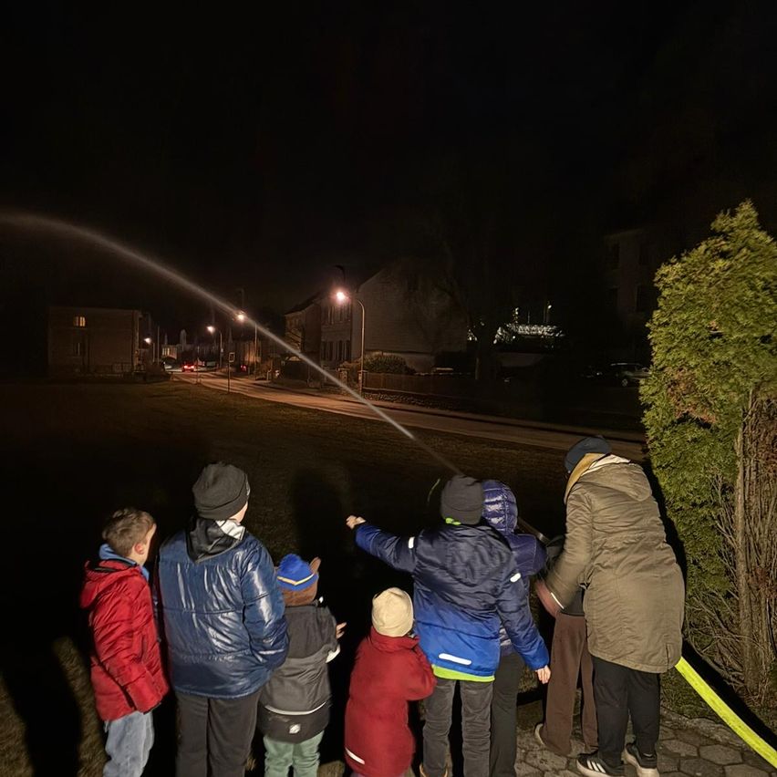 Eine Gruppe von Kindern und Erwachsenen beobachtet eine Wasserfontäne bei Nacht. Die Fontäne spüht Wasser in den dunklen Himmel.