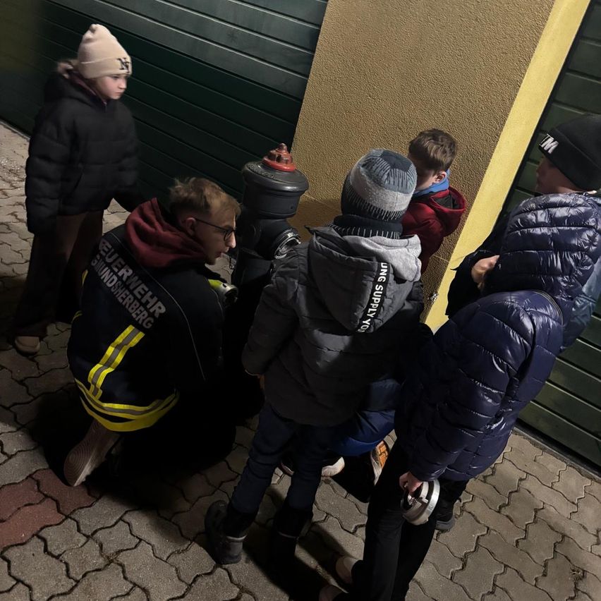 Ein Feuerwehrmann kniet und zeigt etwas einer Gruppe von Kindern auf einem gepflasterten Weg bei Nacht.