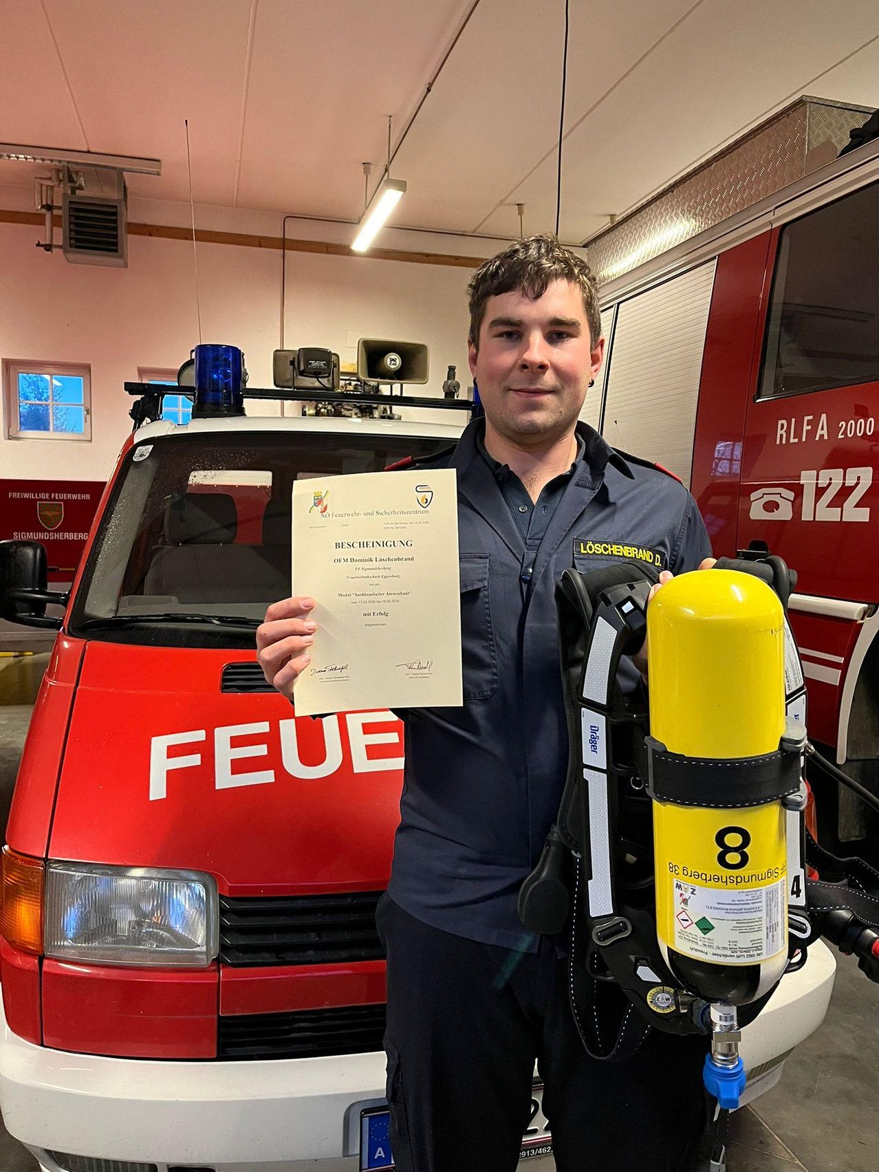 Ein Mann in einer blauen Uniform hält ein Zertifikat und steht neben einem roten Feuerwehrauto mit der Nummer 8 auf einem gelben Feuerlöscher.