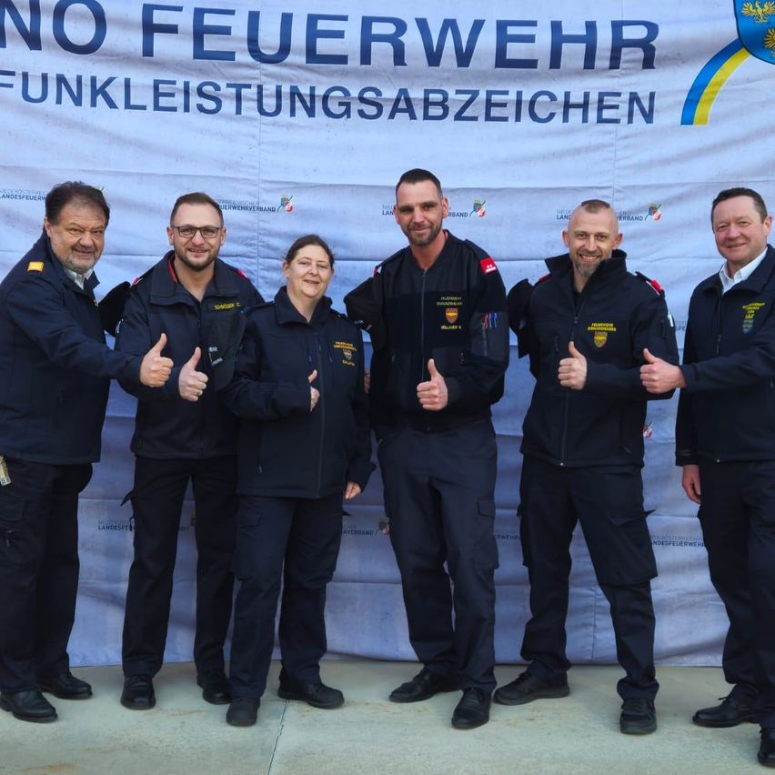 Fünf Feuerwehrleute in Uniform posieren für ein Foto vor einem Banner mit den Worten 'NO FEUERWEHR FUNKLEISTUNSGSABZEICHEN'. Die beiden Frauen und drei Männer lächeln alle und geben ein Daumenhoch.