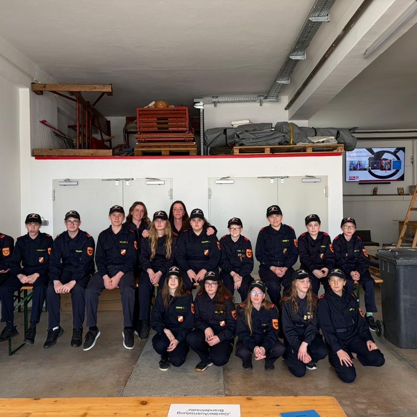 Eine Gruppe Feuerwehrleute in Uniform posiert für ein Foto in einem Raum mit Lagerboxen und einem Monitor.