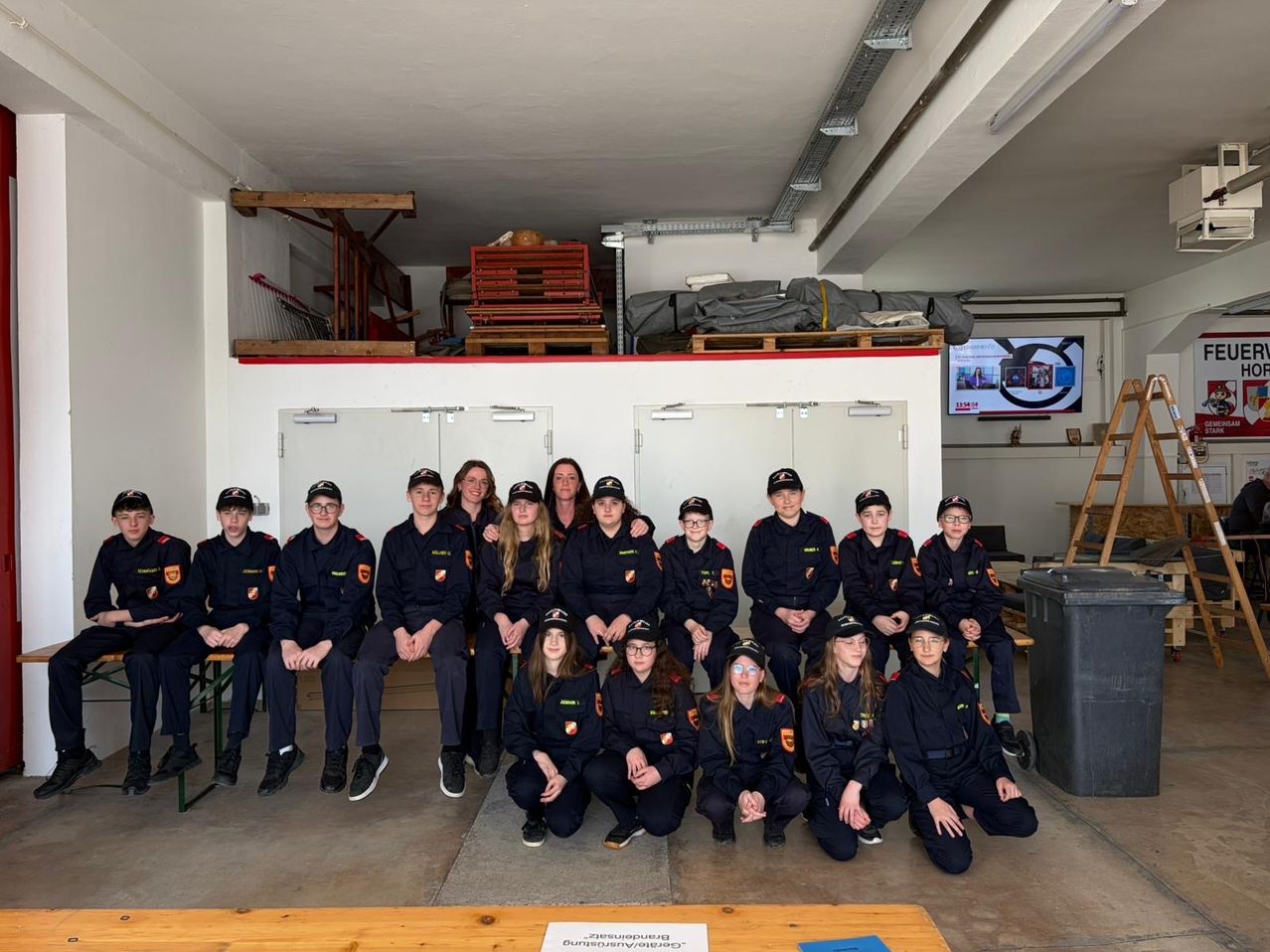 Eine Gruppe Feuerwehrleute in Uniform posiert für ein Foto in einem Raum mit Lagerboxen und einem Monitor.