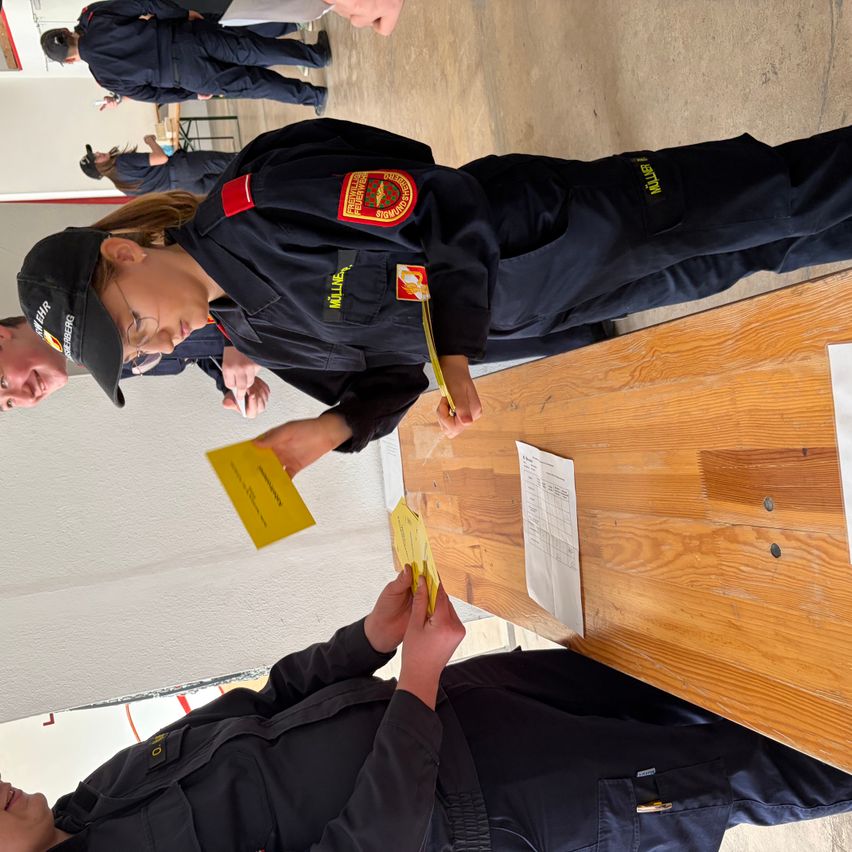 Eine Frau in Uniform hält eine gelbe Karte und steht neben einer anderen Person an einem Holztisch. Dahinter ist teilweise eine Person zu sehen, ebenfalls in Uniform.