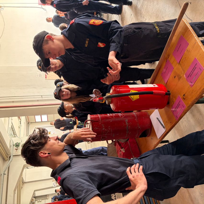 Eine Gruppe von Feuerwehrleuten steht um zwei rote Feuerlöscher herum, möglicherweise während einer Schulung. Eine Person hält den Griff eines Löschers, während eine andere Person versucht, ihn zu berühren.