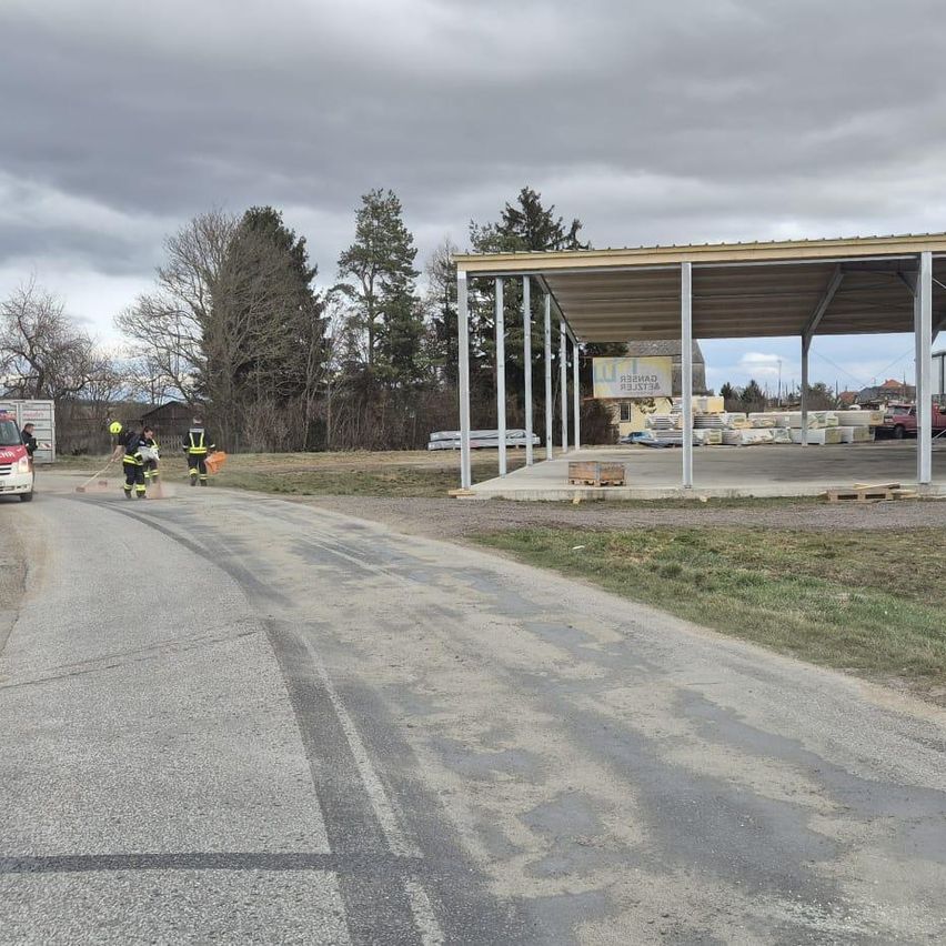 Mehrere Arbeiter in Schutzkleidung reinigen eine Straße, mit einer Baustelle und einem Lagerhaus im Hintergrund. Ein roter LKW ist rechts geparkt.