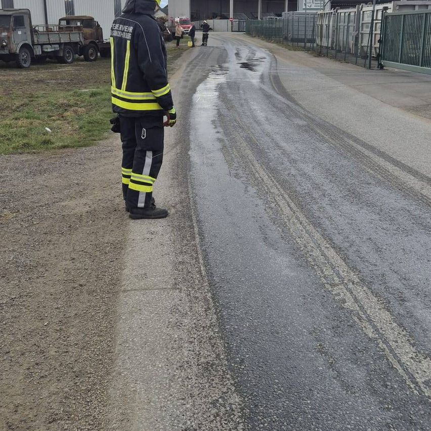 Ein Feuerwehrmann in Uniform steht auf einer nassen Straße und blickt auf geparkte Lastwagen und Menschen. Die Straße ist nass und reflektiert Licht, mit Reifenspuren sichtbar. Es gibt Metallzäune und ein Tor im Hintergrund.