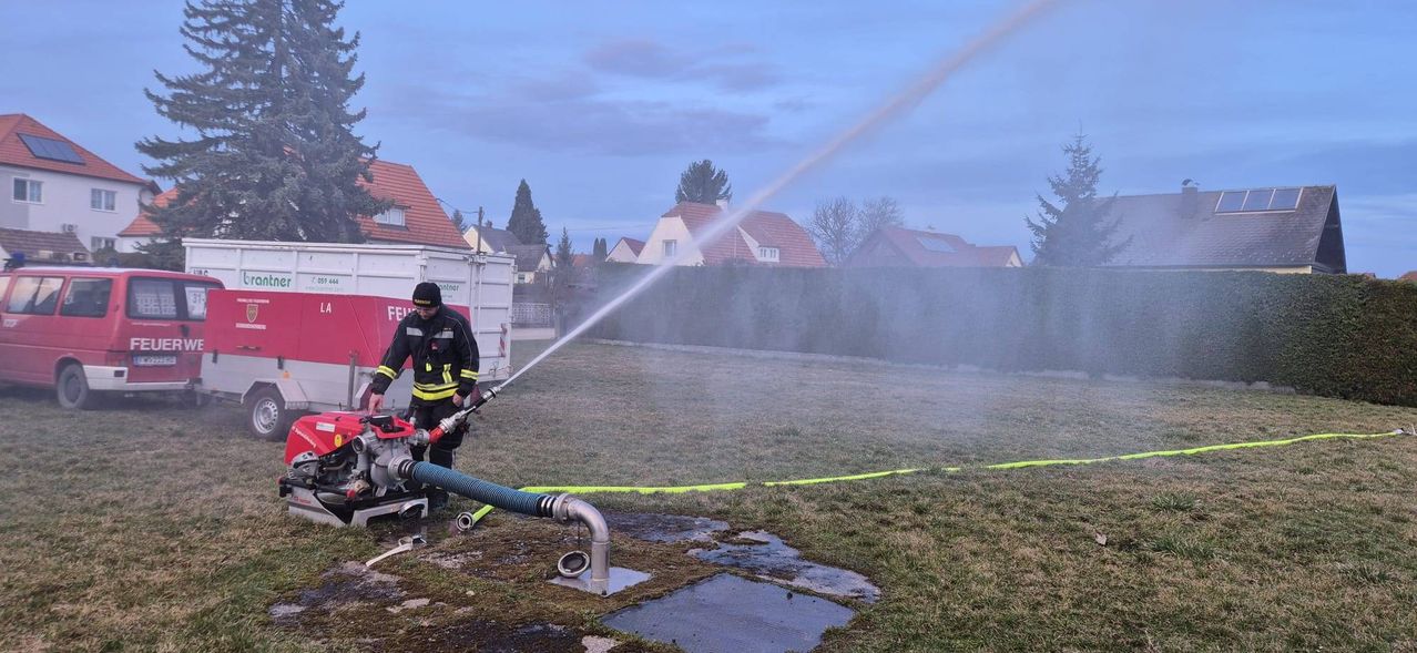 Ein Feuerwehrmann spritzt Wasser aus einem Schlauch, um ein Feuer in der Nähe eines Wohngebiets zu löschen. Der Schlauch ist an einen Wassertank angeschlossen.