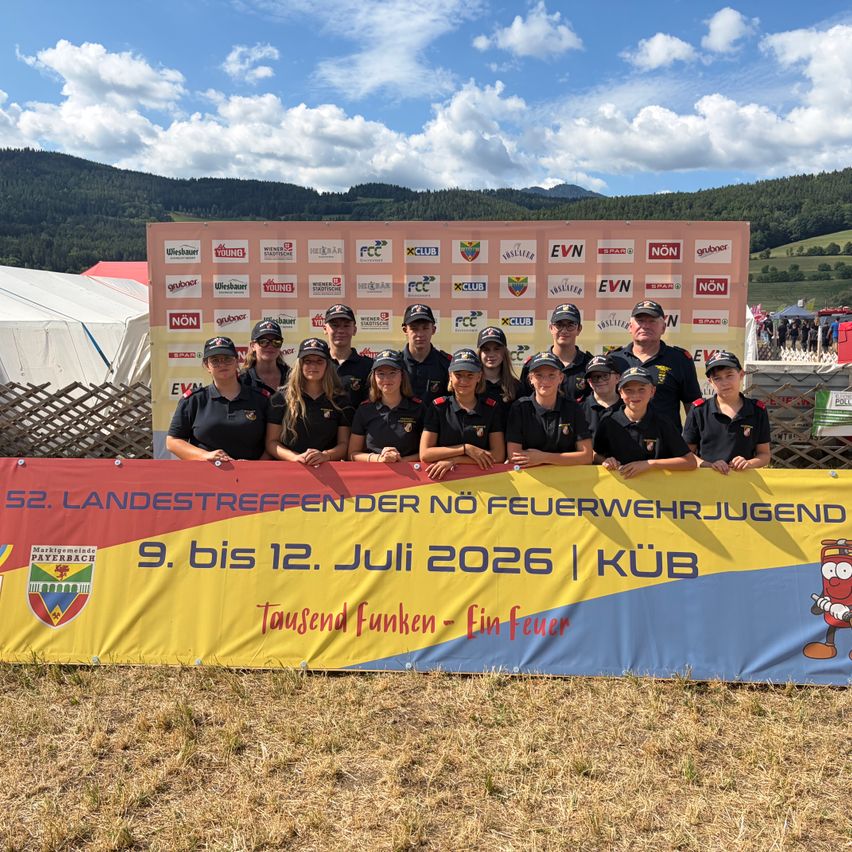 Eine Gruppe junger Feuerwehrleute in Uniform posiert für ein Foto vor einem Banner mit Logos und Bergen im Hintergrund.