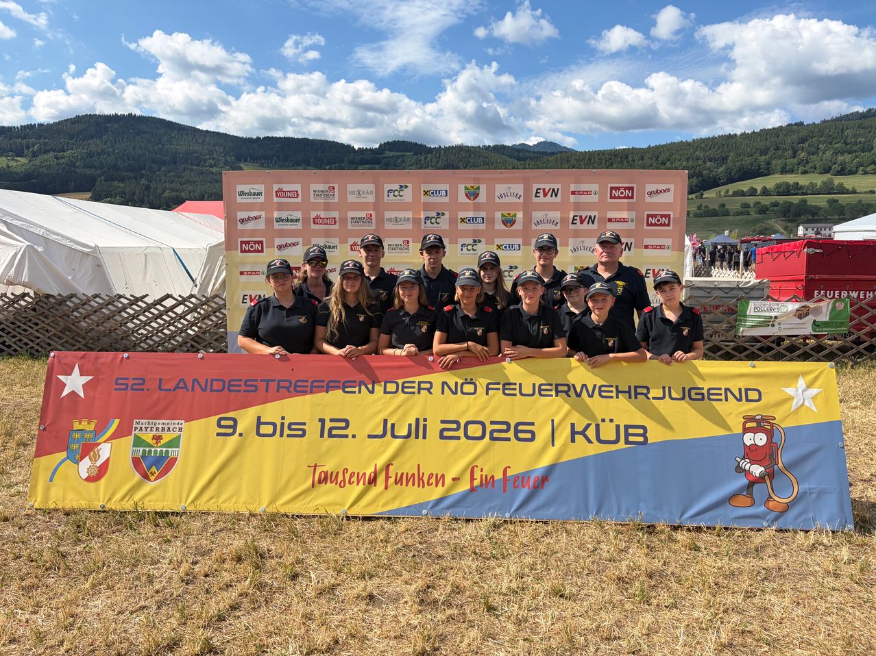 Eine Gruppe junger Feuerwehrleute in Uniform posiert für ein Foto vor einem Banner mit Logos und Bergen im Hintergrund.