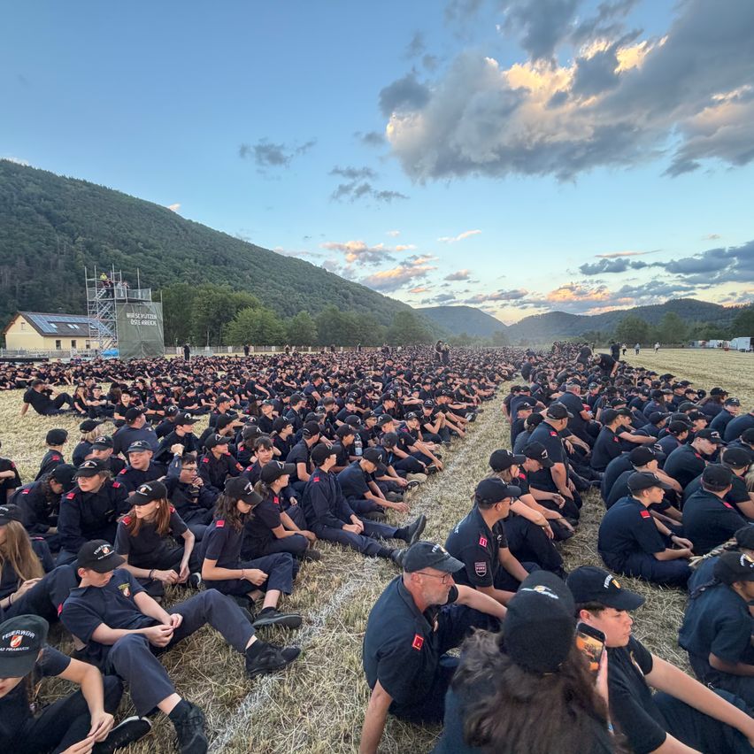 Eine große Gruppe von Menschen in Uniform sitzt auf dem Gras mit Bergen im Hintergrund.
