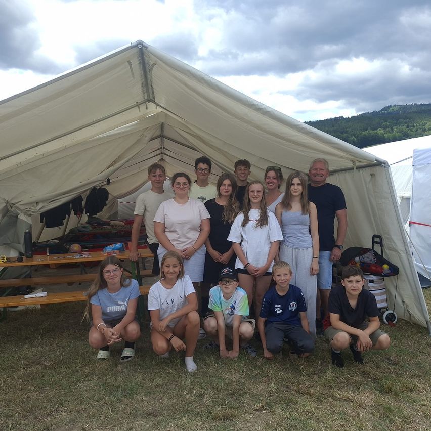 Eine Gruppe von Menschen, einschließlich Kinder und Erwachsene, posiert für ein Foto vor einem großen weißen Zelt, das in einem Grasfeld aufgebaut ist.