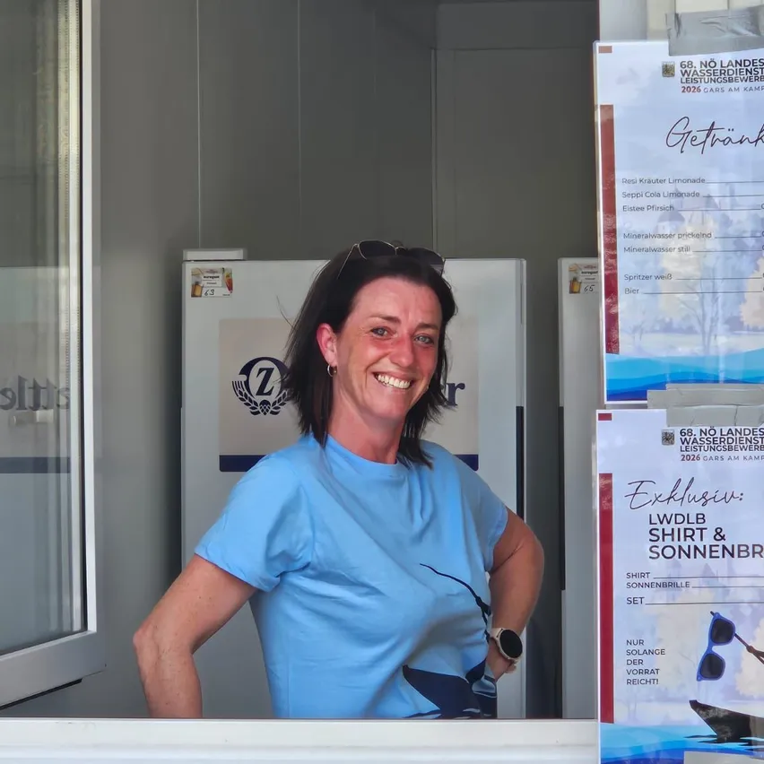 Eine Frau mit Sonnenbrille auf dem Kopf und einem blauen T-Shirt lächelt vor einer Glastür mit der Aufschrift '7'. Hinter ihr steht ein Kühlschrank mit einem Etikett und zwei Postern an der Wand.