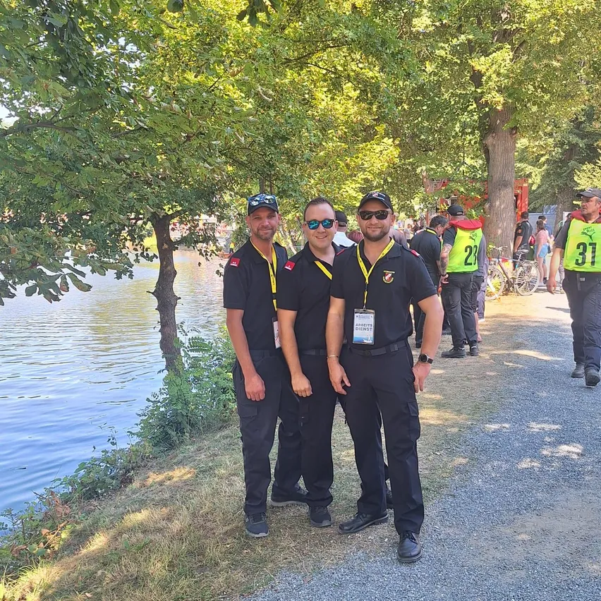 Drei Männer in Uniform stehen nebeneinander am See, mit Bäumen und einem Kiesweg im Hintergrund.