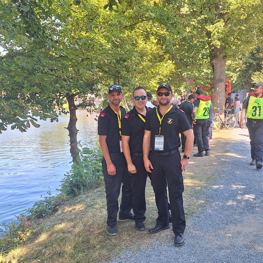 Drei Männer in Uniform stehen nebeneinander am See, mit Bäumen und einem Kiesweg im Hintergrund.