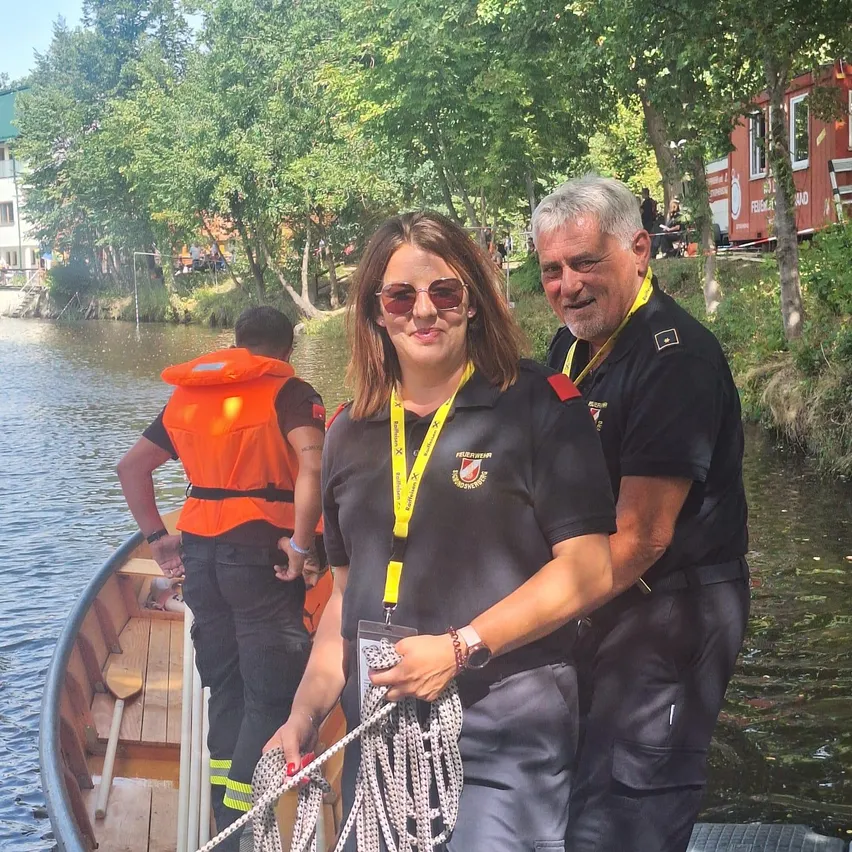 Eine Frau mit einem gelben Ausweisband steht neben zwei Männern in schwarzen Uniformen auf einem Boot und hält Seile, mit einem Mann in einem orangefarbenen Rettungsweste im Hintergrund.