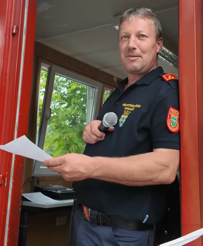 Ein Mann in Uniform steht in einem Raum, hält ein Mikrofon in der Hand und liest von einem Blatt Papier. Der Raum hat eine rote Tür und ein Fenster mit Bäumen draußen.