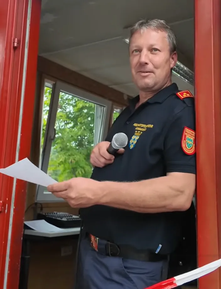 Ein Mann in Uniform steht in einem Raum, hält ein Mikrofon in der Hand und liest von einem Blatt Papier. Der Raum hat eine rote Tür und ein Fenster mit Bäumen draußen.