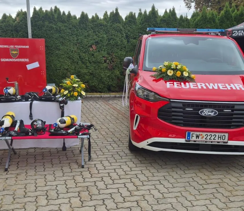 Ein rotes Ford-Fahrzeug mit dem Wort 'Feuerwehr' auf der Front, geparkt auf einer Ziegelfläche. In der Nähe ein Tisch mit Tauchausrüstung und Blumen.
