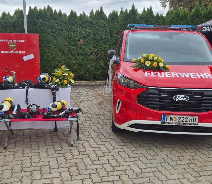 A red Ford vehicle with the word 'Feuerwehr' on the front, parked on a brick surface. Nearby, a table with diving equipment and flowers.