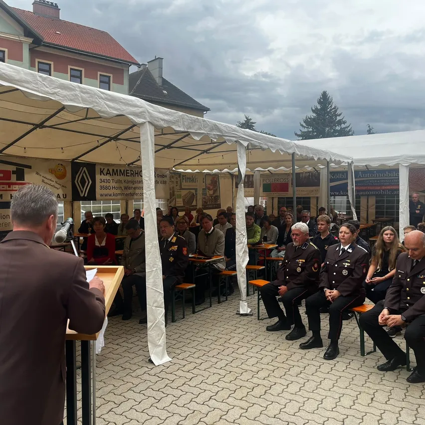 Eine formelle Versammlung unter weißen Zelten mit einem Mann, der an einem Podium spricht, umgeben von uniformierten Teilnehmern.
