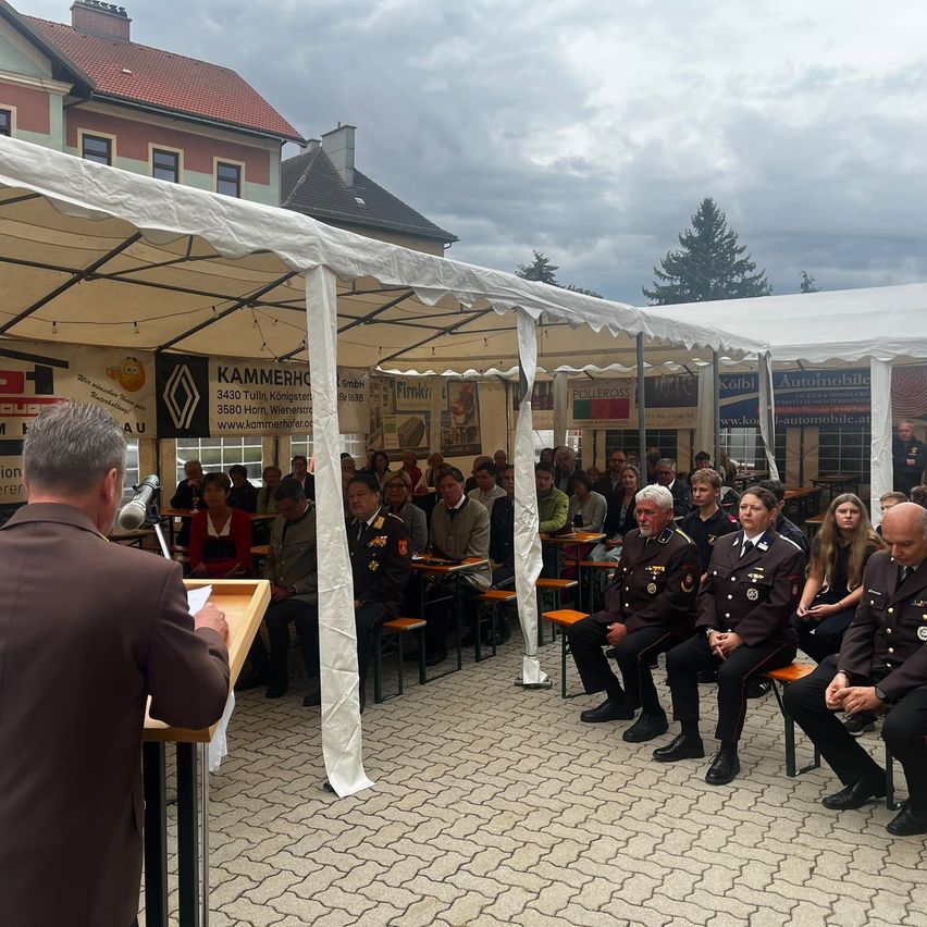 A formal gathering under white tents with a man speaking at a podium, surrounded by uniformed attendees.
