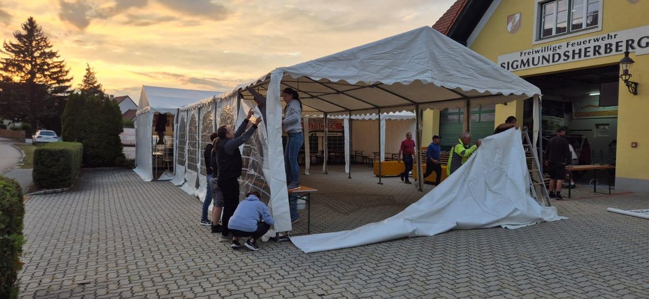 A group of people are setting up a large white tent with a metal frame. One person is standing on a table, while others are assisting. The tent is in an outdoor area with brick flooring.