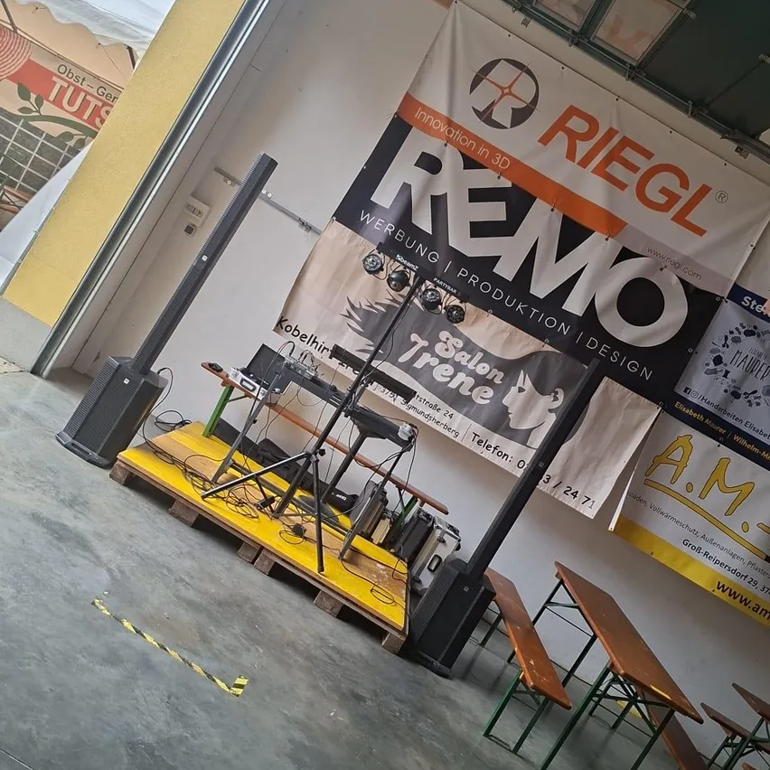 Ein Bühnenaufbau mit Lautsprechern, einem Keyboard und einem Banner, das 'Riegl' und 'Remo' bewirbt. Die Bühne steht auf einer gelben Plattform mit einem Stativ in der Mitte. Im Hintergrund befinden sich Bänke und Tische.