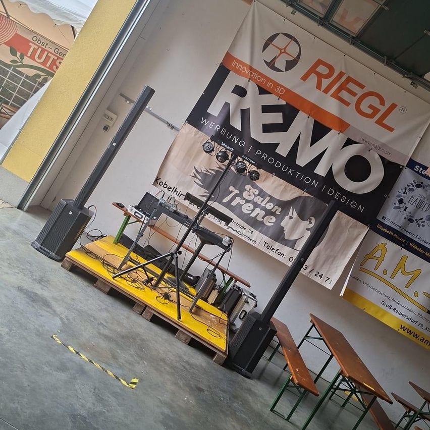 Ein Bühnenaufbau mit Lautsprechern, einem Keyboard und einem Banner, das 'Riegl' und 'Remo' bewirbt. Die Bühne steht auf einer gelben Plattform mit einem Stativ in der Mitte. Im Hintergrund befinden sich Bänke und Tische.