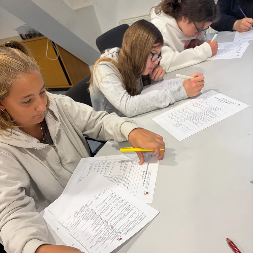 Drei junge Frauen sitzen an einem Tisch und konzentrieren sich jeweils auf ein Blatt Papier mit einem Stift in der Hand. Die Frau im Vordergrund hält einen gelben Stift.