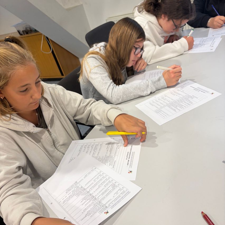 Drei junge Frauen sitzen an einem Tisch und konzentrieren sich jeweils auf ein Blatt Papier mit einem Stift in der Hand. Die Frau im Vordergrund hält einen gelben Stift.
