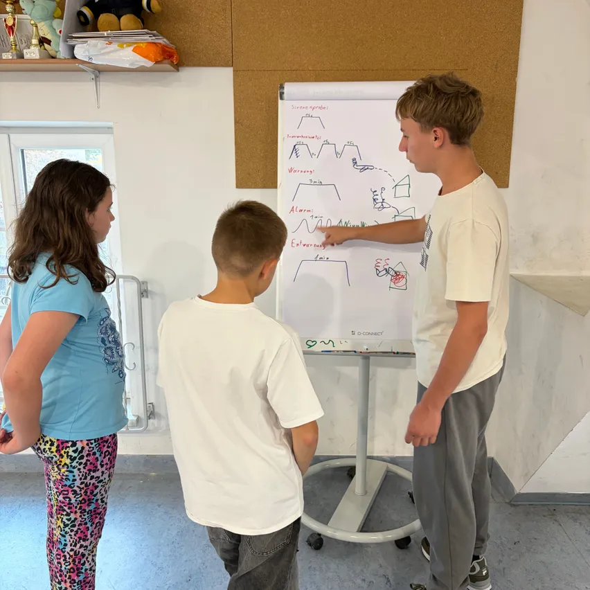 Drei Schüler stehen um eine Tafel in einem Klassenzimmer, einer der Jungen zeigt auf die Tafel. Eines Mädchen in einem blauen Shirt und bunten Hosen steht links, ein anderer Junge in einem weißen Shirt steht in der Mitte.
