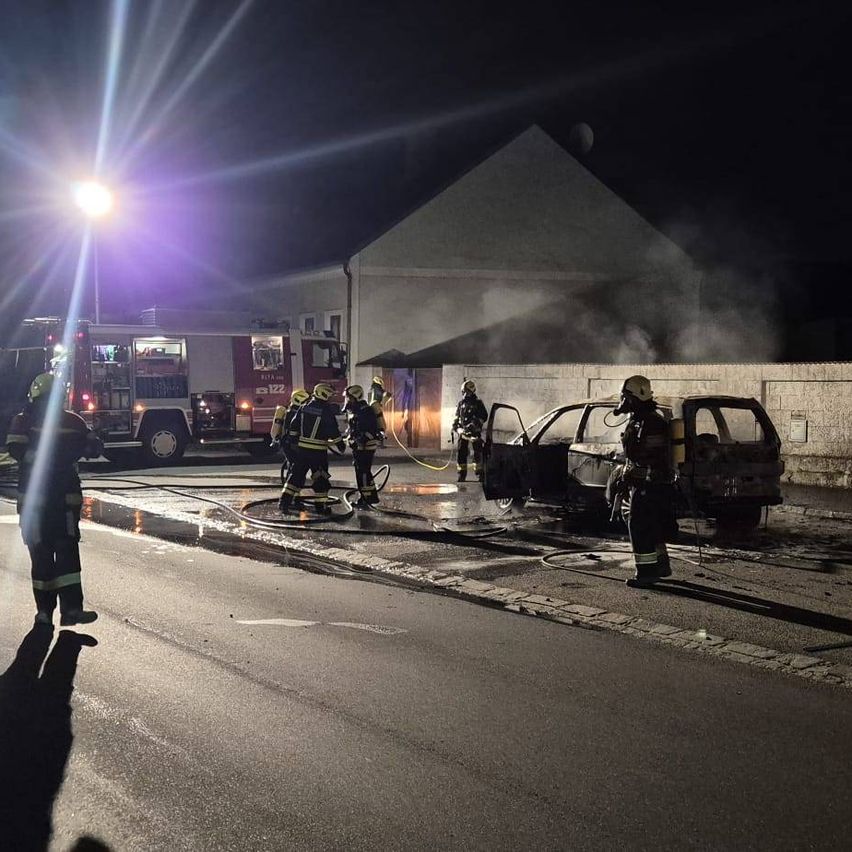 Feuerwehrleute löschen ein brennendes Auto bei Nacht. Das Fahrzeug steht mit Rauch und Feuerwehrleuten darum auf der Straße.