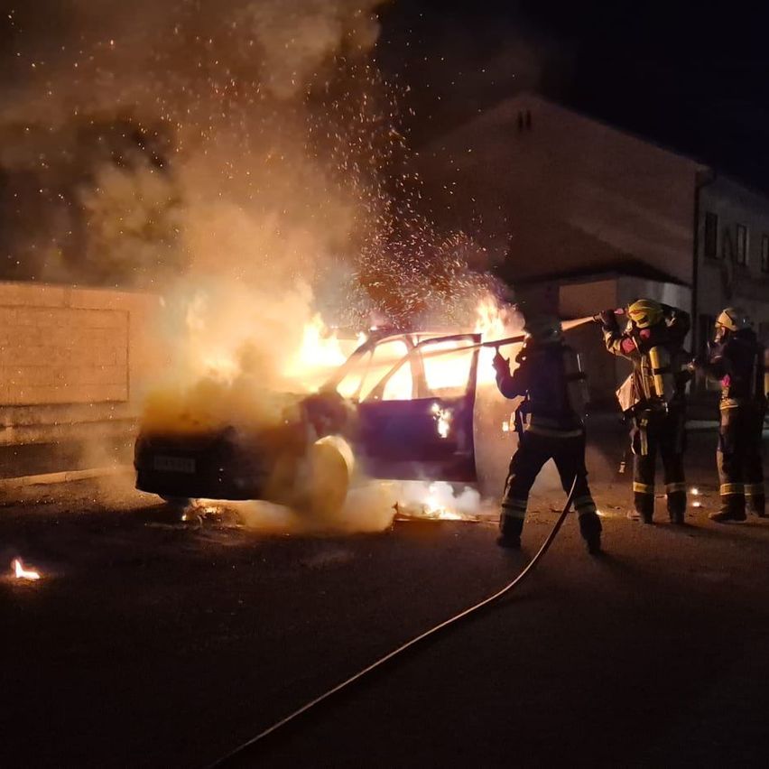 Feuerwehrleute löschen ein brennendes Auto in der Nacht auf der Straße. Flammen und Rauch erfüllen die Luft, zwei Feuerwehrleute benutzen Schläuche.