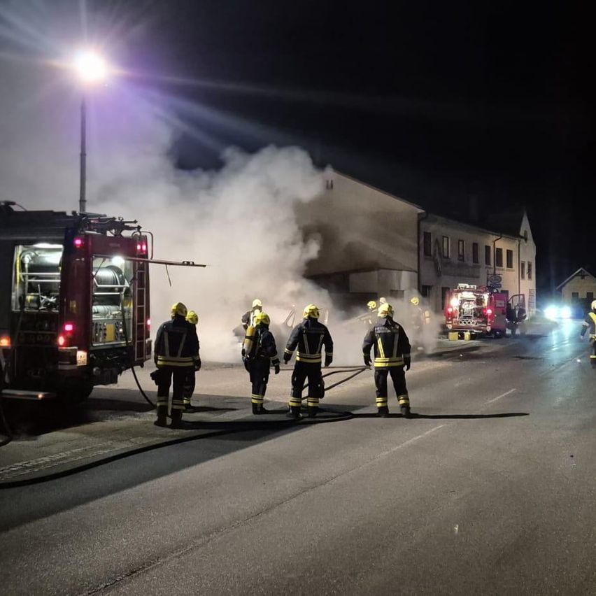 Feuerwehrleute stehen nachts auf einer Straße mit Rauch und einem Feuerwehrwagen im Hintergrund.