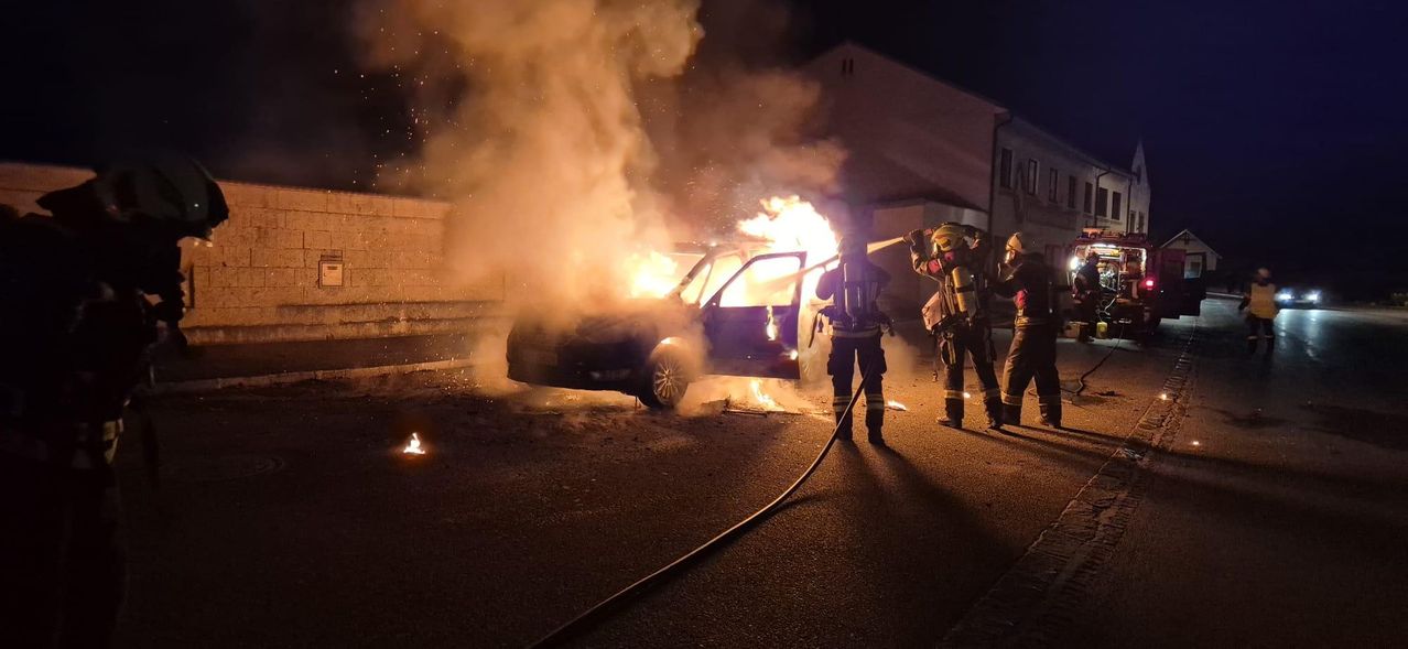 Feuerwehrleute in Schutzausrüstung löschen ein brennendes Auto. Rauch und Flammen steigen aus dem Fahrzeug auf.
