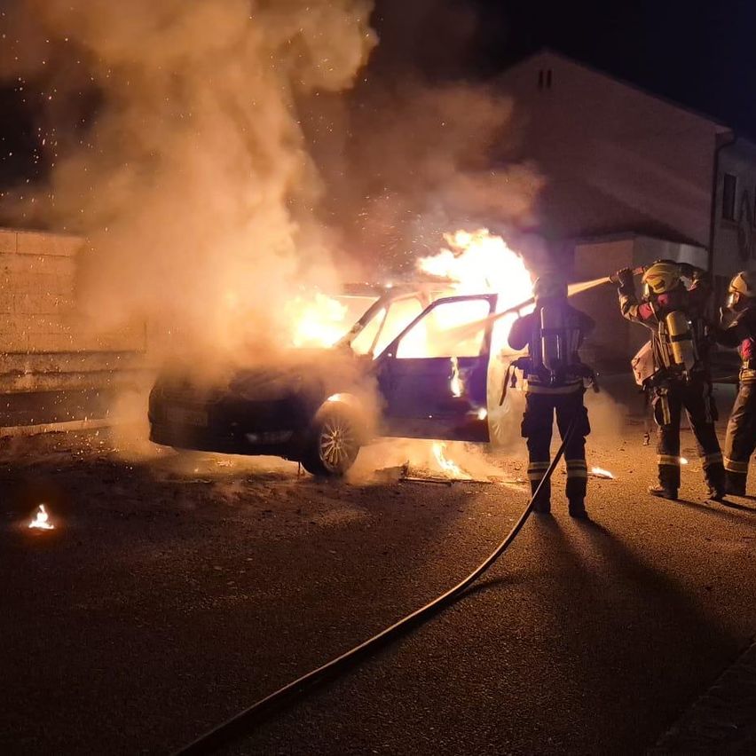 Feuerwehrleute in Schutzausrüstung löschen ein brennendes Auto. Rauch und Flammen steigen aus dem Fahrzeug auf.
