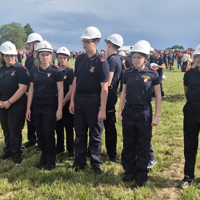 Eine Gruppe uniformierter Personen steht in einer Reihe auf einem Rasenfeld. Sie tragen Helme und Uniformen mit Abzeichen. Im Hintergrund versammeln sich weitere Menschen, einige mit Hüten.