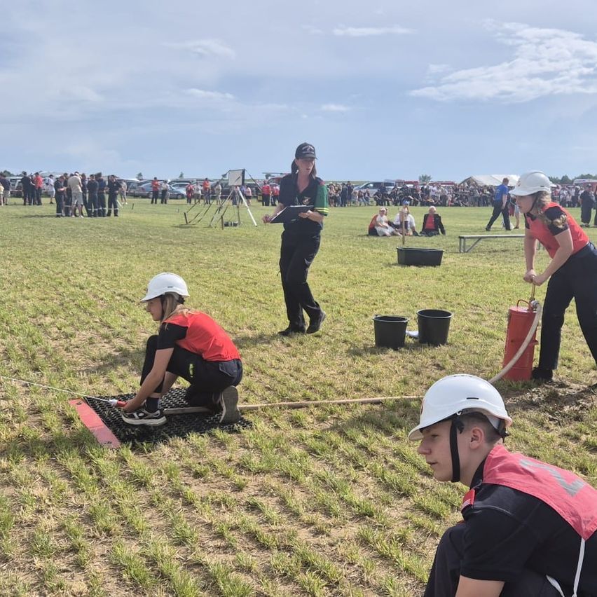 Eine Gruppe von Personen in roten und schwarzen Uniformen führt eine Aufgabe auf einem Rasenfeld unter einem bewölkten Himmel aus. Zwei halten Seile, einer hockt auf einem Matten, und einer steht mit einem Klemmbrett. Zuschauer beobachten im Hintergrund.