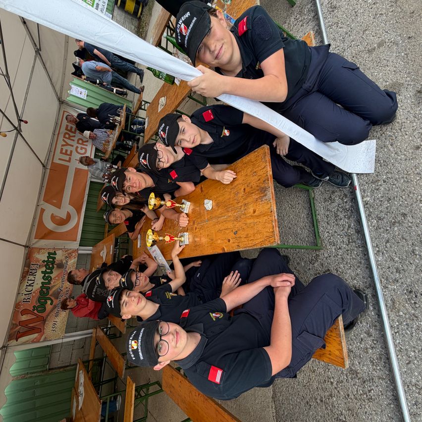 Eine Gruppe von Menschen in schwarzen Uniformen, wahrscheinlich Feuerwehrleute, sitzen auf Bänken um einen Tisch mit Auszeichnungen herum. Sie lächeln und posieren für ein Foto.