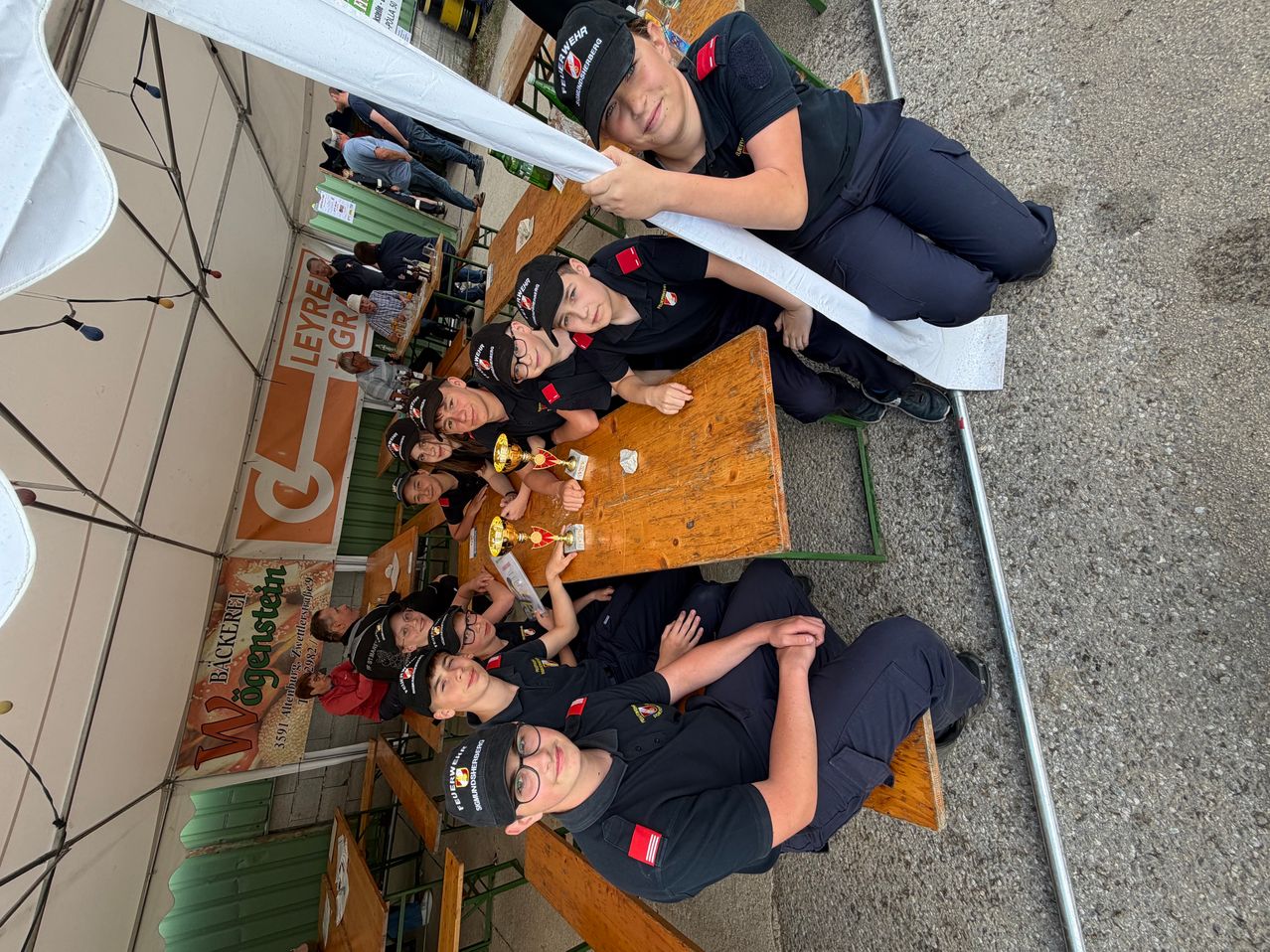 Eine Gruppe von Menschen in schwarzen Uniformen, wahrscheinlich Feuerwehrleute, sitzen auf Bänken um einen Tisch mit Auszeichnungen herum. Sie lächeln und posieren für ein Foto.
