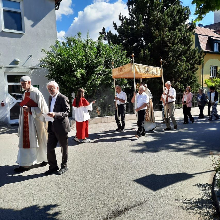 Priester und Menschen bei einer Prozession auf einer Straße, mit einem Baldachin, Bäumen und Gebäuden im Hintergrund.