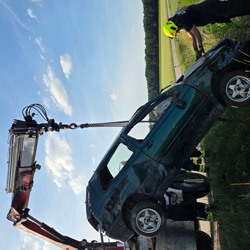 Ein grünes Fahrzeug wird von einem Kran vom Boden angehoben. Eine Person mit gelbem Helm steht an der Seite des Fahrzeugs.