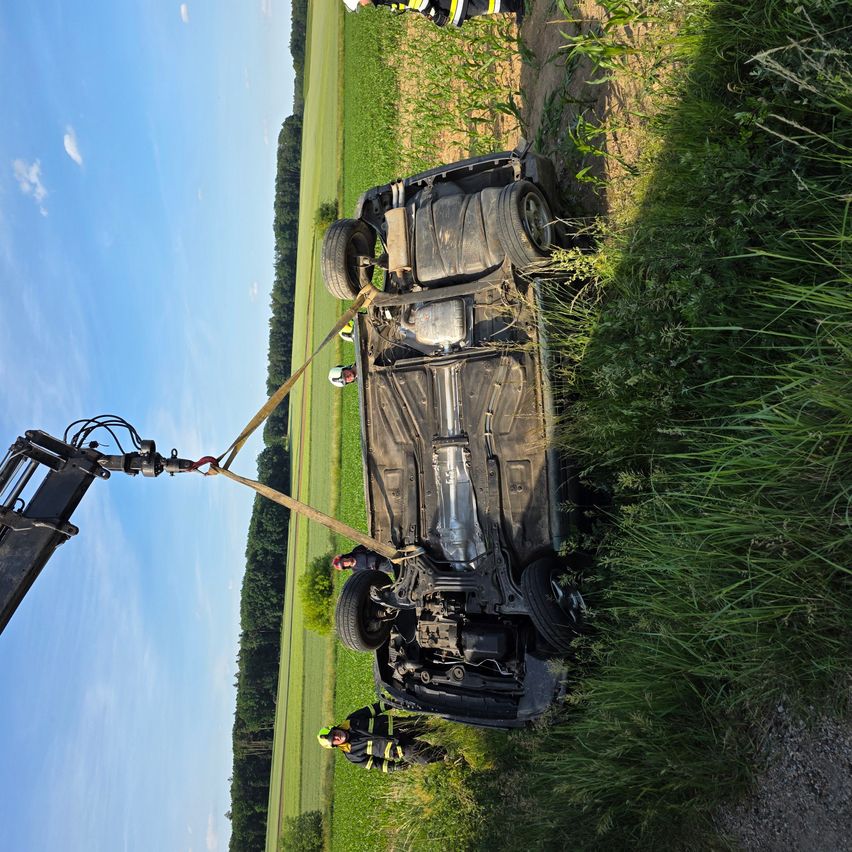 Ein demoliertes Fahrzeug wird von einem Kran aus einem Grasfeld gehoben, in der Nähe sind Arbeiter.
