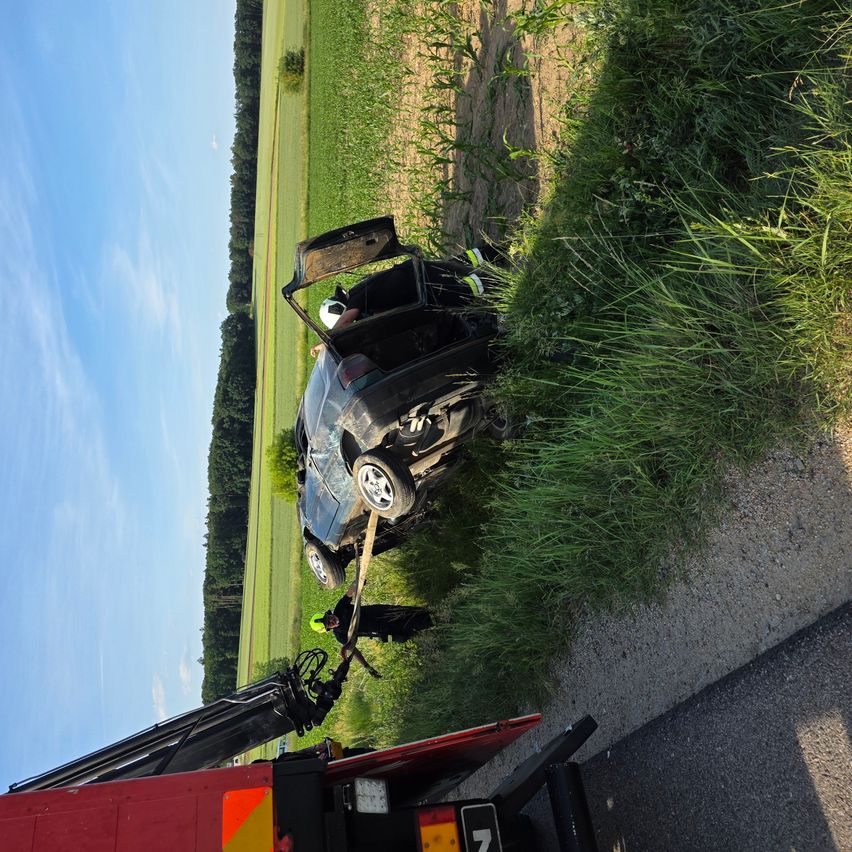 Ein demoliertes Auto wird von einem Abschleppwagen an einem Feldrand unter einem blauen Himmel gezogen.