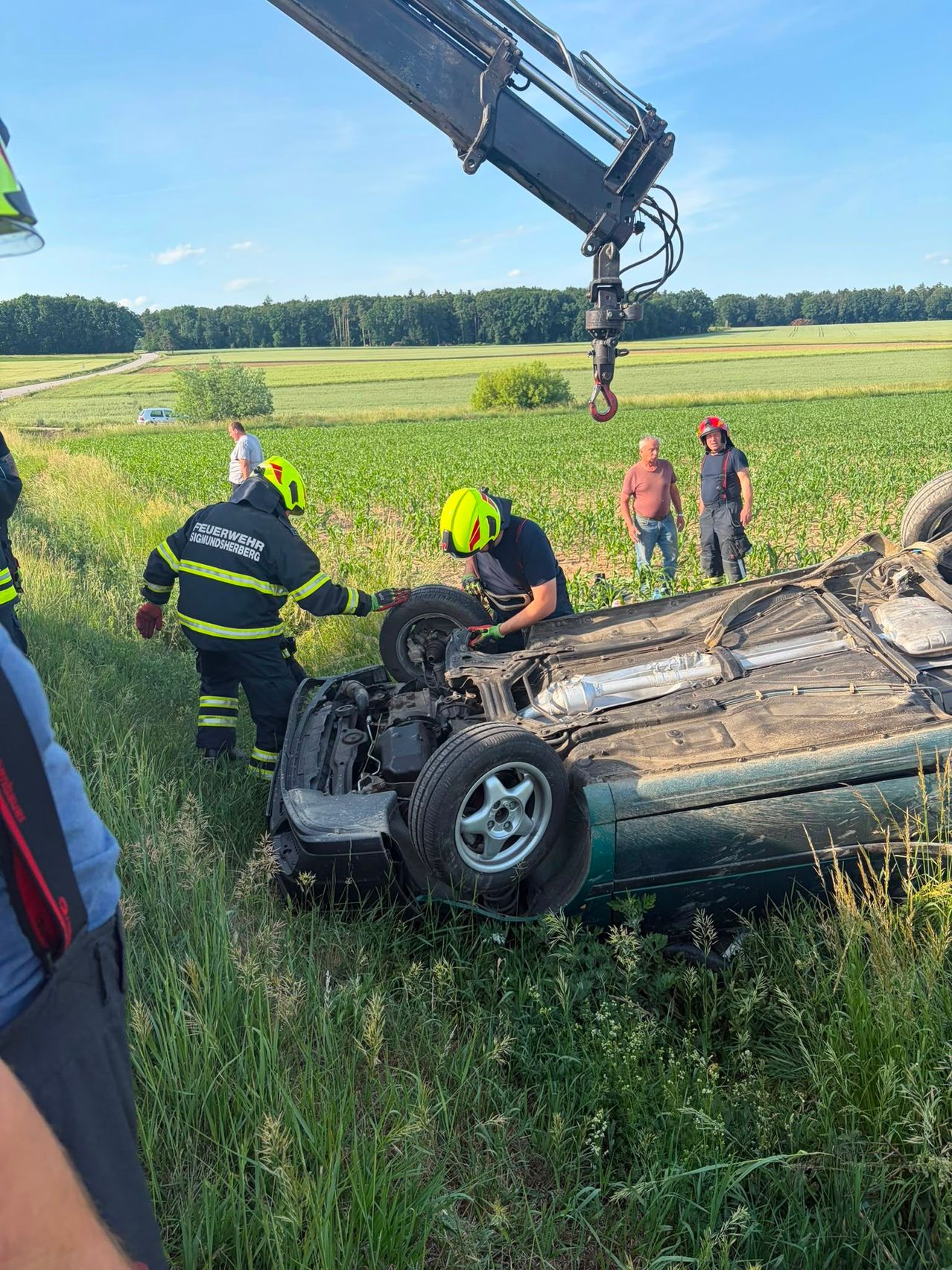Feuerwehrleute untersuchen ein umgekipptes Auto auf einem Grasfeld. Zwei Personen mit Helmen arbeiten am Motor des Fahrzeugs. Ein Mann im weißen Hemd und ein Mann mit rotem Helm stehen in der Nähe.