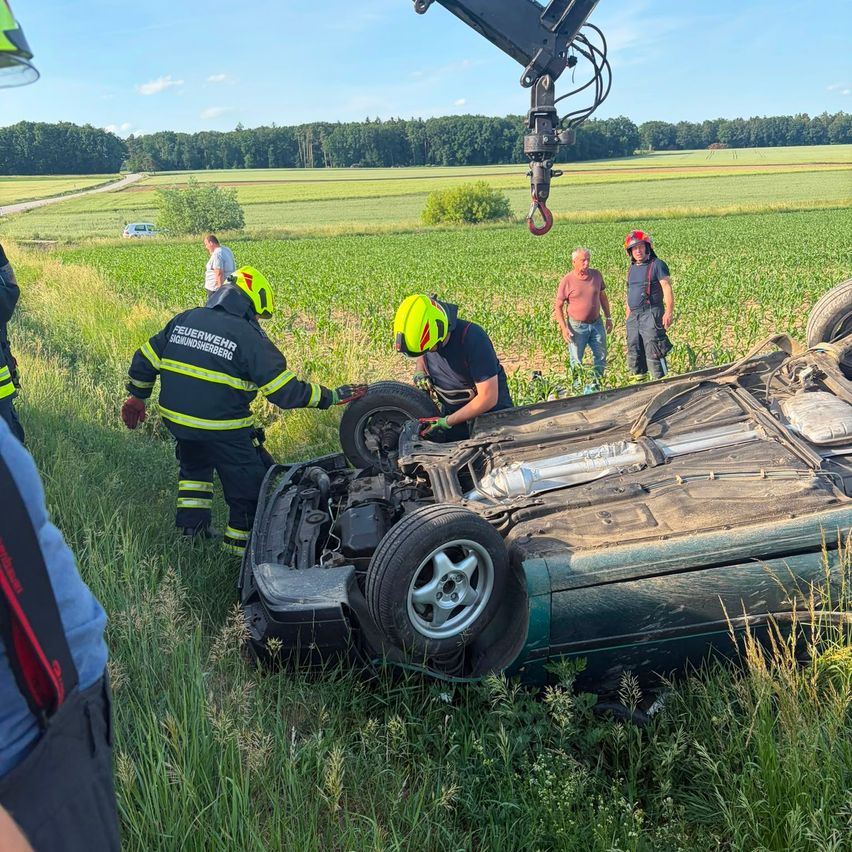 Feuerwehrleute untersuchen ein umgekipptes Auto auf einem Grasfeld. Zwei Personen mit Helmen arbeiten am Motor des Fahrzeugs. Ein Mann im weißen Hemd und ein Mann mit rotem Helm stehen in der Nähe.